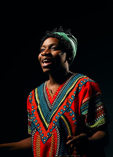 Against a black background a woman in a red, blue, black, green and yellow patterned top, in an African print, with a green hair-wrap, is looking off to the left of the image and smiling/singing.