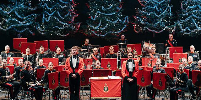 The Band of HM Royal Marines stand on the Festival Theatre Stage dressed in red military jackets, facing out front. Behind them are five blue-lit Christmas Trees.