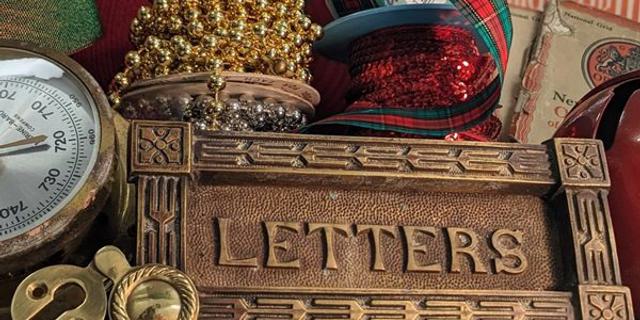 A brass letter box sign is surrounded by festive ribbons and beads in red, green and gold. Booklets of Christmas carols and maps of Chichester as well as locks and atmospheric dials also are there all on a silver glittery backdrop.