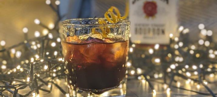 A Winter's night cocktail. A short glass filled with ice and dark colour liquid. Fairy lights sit behind and a bottle of Jim Beam behind.