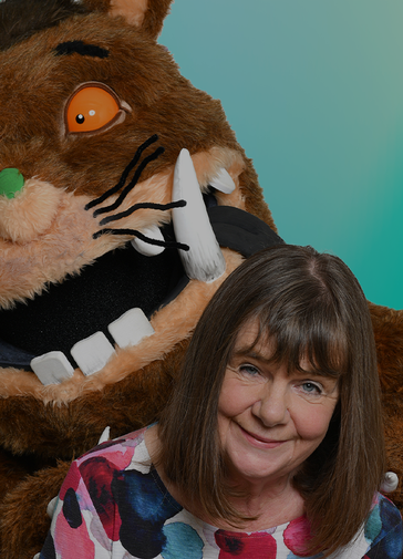 Foreground right of the image is Julia Donaldson in a white top with blue, pink and green splotches on it. Behind her is a large brown furry gruffalo with white tusks, black whiskers, a green nose and orange eyes. The background is turquoise.