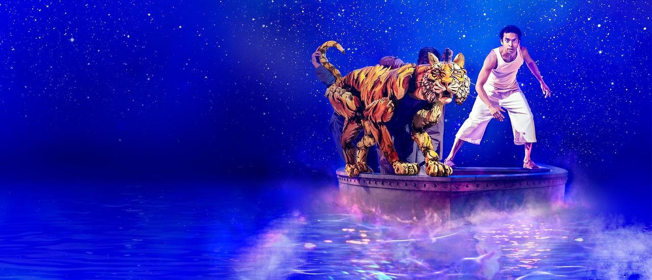 A large bengal tiger puppet and a young man dressed in a white vest and cropped white trousers are both crouched on the bow of a small lifeboat, staring out at the camera. Behind them is a vivid blue sky scattered with stars, which is reflected in the glowing sea beneath the boat.