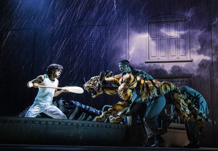 A dark stage can be seen, with projections of rain and lightning. A young man wearing white holds an oar as a weapon. He is facing off with a roaring puppet tiger. The tiger is lunging aggressively, puppetered by three people.