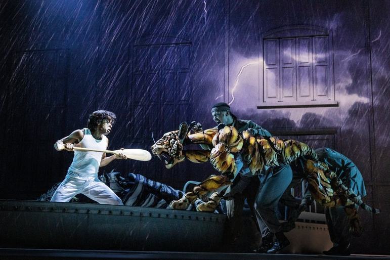 A dark stage can be seen, with projections of rain and lightning. A young man wearing white holds an oar as a weapon. He is facing off with a roaring puppet tiger. The tiger is lunging aggressively, puppetered by three people.