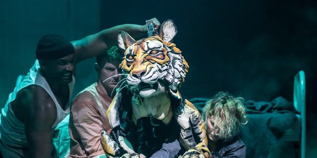In the centre of the frame is a puppet orange and white tiger, with three puppeteers controlling its legs, head and body. The puppetters are looking intently at the tiger. In the background is a metal bed frame.