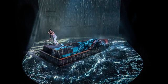 A wide view of a boat on a stage, with projections of blue and grey rough water and rain all around it. A young man in a white vest and trousers stands on to the boat, with his mouth and eyes wide in panic. A puppet tiger is lying on the boat next to him.