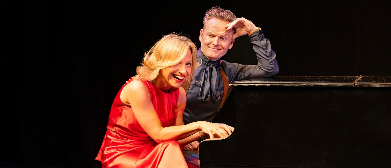 Liza Pulman & Joe Stilgoe sit at a black grand piano; she is wearing a long orange dress and orange platform shoes; he;s wearing a grey shirt and grey silky tie. She is laughing; he is smiling and looking at her.