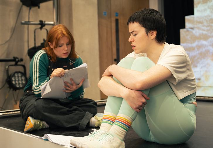 Two performers  sit on the floor of a rehearsal studio. The performer  on the left has shoulder length ginger hair and wears a green sweatshirt under black dungarees with yellow trainers. They are holding a script and reading from it. The performer  on the right is looking down at another script on the floor by their feet.  They have short dark hair and  wear  matching sage green leggings and a crop top with a cropped white t-shirt on top, rainbow stripey socks and white trainers.