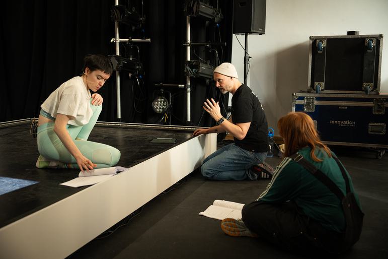 Two performers  and the director sit on the floor of a rehearsal studio. The two performers  look down at scripts  and the director has a tablet in front of him. The actor on the left sits up on a low stage, with one knee hugged to their chest. They are concentrating on the script. The other performer  has their back to the camera, looking at the director, who sits in between the two. He is looking intently at the performer  in front of him and speaking to them, with one hand lifted.