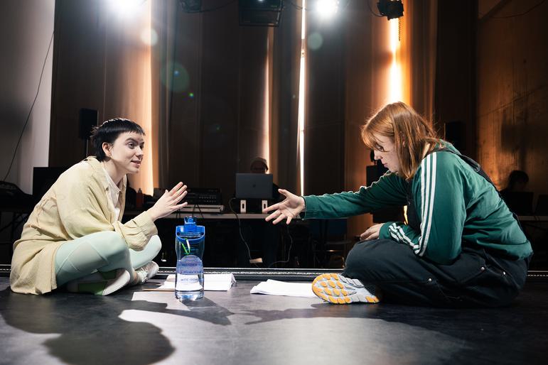 Two performers sit on the floor of a rehearsal studio, side on to the camera. The performer on the left has short dark hair and wears matching sage green leggings and a crop top with a cropped white t-shirt and checked yellow shirt on top, rainbow stripey socks and white trainers. They are looking ahead and speaking with one hand lifted. The performer on the right has shoulder length ginger hair and wears a green sweatshirt under black dungarees with yellow trainers. They hold their arm outstretched and look down at a script on the floor.