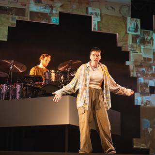 A performer stands centre stage, wearing khaki trousers, a light coloured top and a checked shirt. They have their arms out, as though questioning, talking out to the audience inquisitively. Behind them is a drummer, playing the drums. There is a geometric arch around them with images projected all over it.