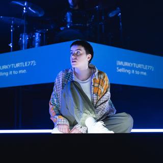 A performer sits on the stage, looking off to the left with a distant, thoughtul expression. They wear khaki trousers, a light coloured top and a checked shirt with white trainers. Behind them a blue strip is projected, which reads 'MurkyTurtle77: Selling it to me.'. A drum kit can be seen in the background.