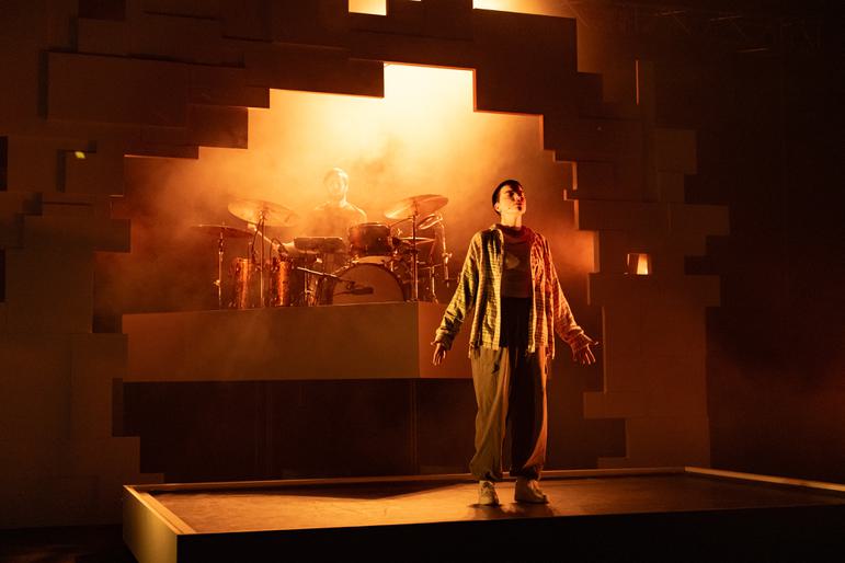 A stage is lit with orange and yellow lights and filled with smoky haze. A performer stands centre stage wearing baggy trousers, trainers and a checked shirt. They have their arms by their sides and their head tilted back with their eyes closed, as though praying or meditating. There is a geometric archway behind them and a drummer playing the drums.