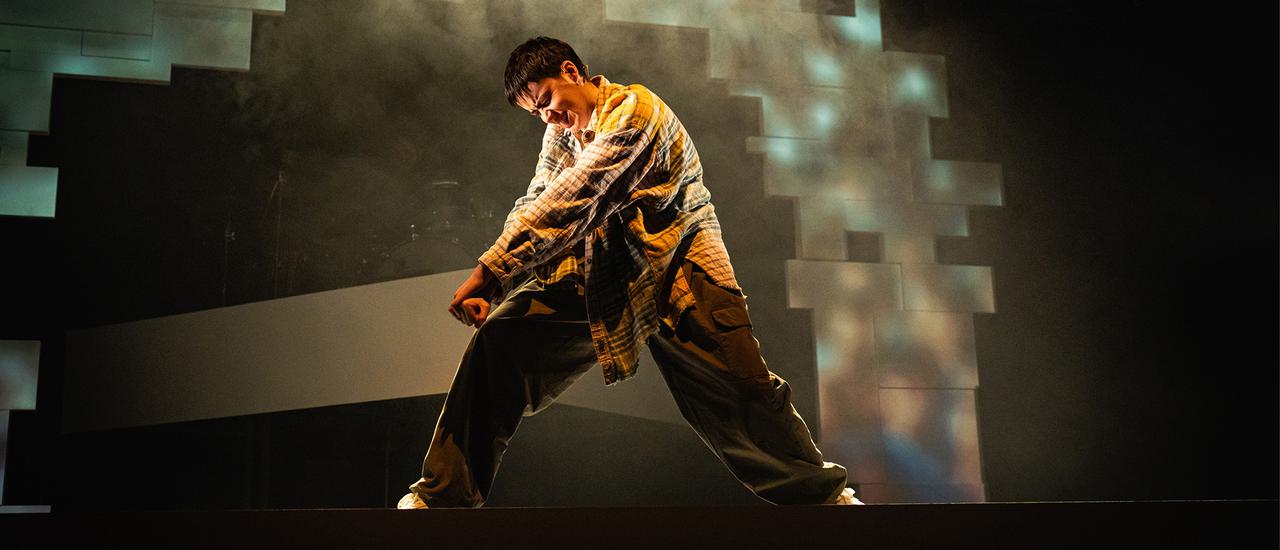 A performer stands lunging to the side with legs wide, arms in fists pointing down towards the front leg. They are closing their eyes tight, like they are concentrating hard. Behind them there is a white geometric archway, lit in bluey grey with a mist of stage haze covering it. The stage around that is dark.