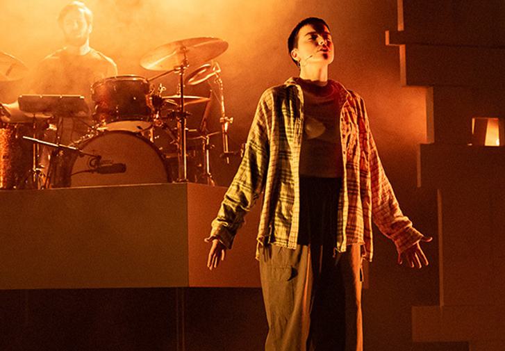 A stage is lit with orange and yellow lights and filled with smoky haze. A performer stands centre stage wearing baggy trousers, trainers and a checked shirt. They have their arms by their sides and their head tilted back with their eyes closed, as though praying or meditating. There is a geometric archway behind them and a drummer playing the drums.