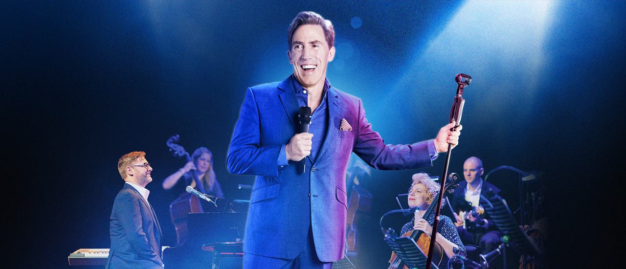 Rob Brydon is spotlighted holding a microphone and smiling broadly. He's wearing a dark suit and standing in front of five musicians seated behind him.