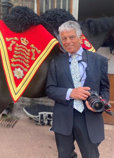 Photographer Henry Dallal stands next to the King's horse (which has a bright red formal dressing on this neck). Henry is dressed in a dark blue suit and has a camera in his hands.