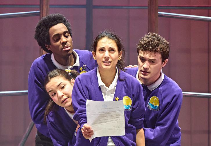 Four students in matching purple school uniforms stand huddled together on the stage. They are all looking with interest at a piece of paper that the actor in the centre is holding. She is looking up at the audience and speaking, looking determined, with her fist clenched.
