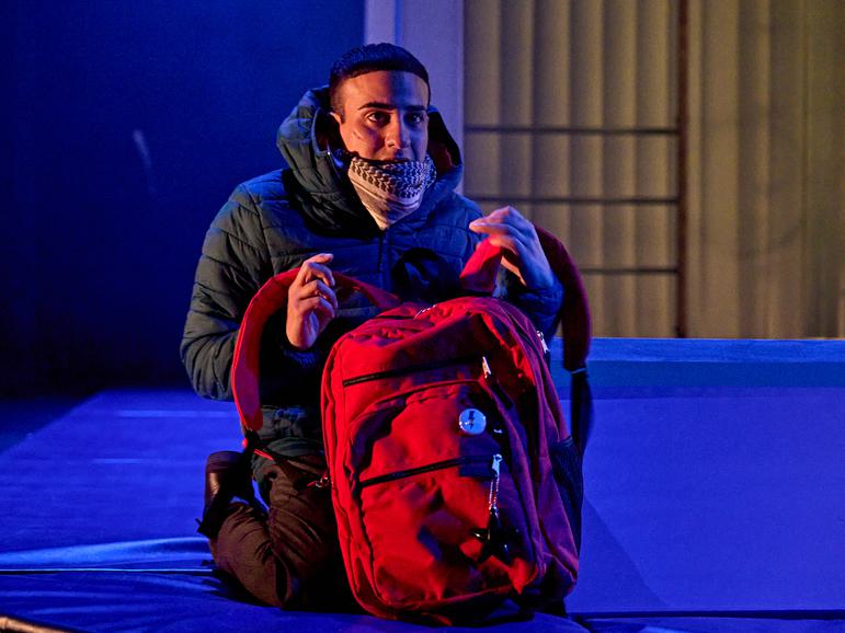 A boy wearing a black puffer jacket and patterned scarf crouches on the stage, which is lit in blue. He looks off to one side, with a worried facial expression. He clutches a big red backpack by the straps.
