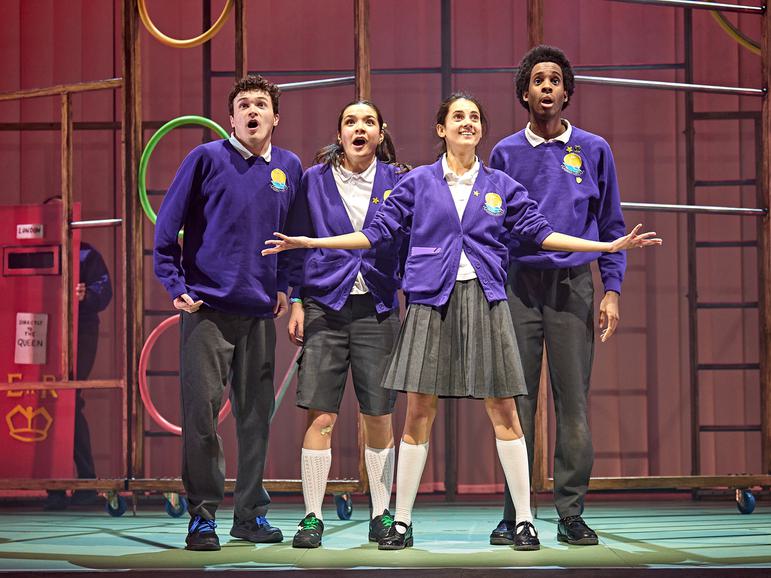 Four students wearing matching purple school uniforms stand together, centre stage. Behind them is a school gymnasium style climbing frame. They all look out at the audience in amazement and one girl in the centre holds her arms out and smiles, like she is showing them something amazing.