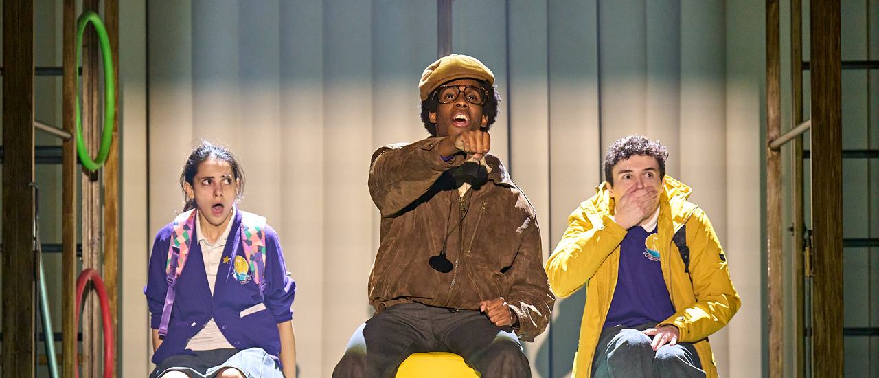 A man wearing a brown aviator jacket and flat cap sits on a yellow wheelie chair, with his arm outstretched, mimicking driving a car. On either side of him sit two students in purple school uniforms, looking shocked; one has her mouth open wide and looks at the man and the other has his hand over his mouth and looks out in front.