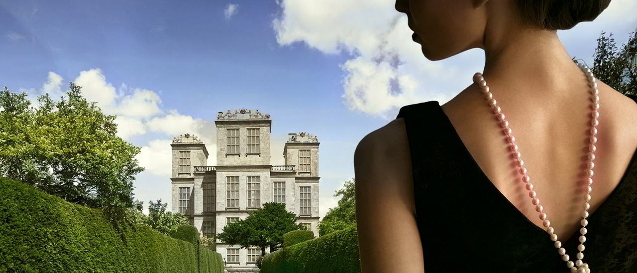 Blue sky with white clouds are the backdrop to a stately home, with green manicured lawns stretching out in front, at the foreground of the image. To the right, up close, is a woman side on, wearing a black dress and black long sleeved gloves, with pearls around her neck.