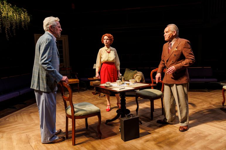 Three older people in smart clothes stand on a parquet wooden floored stage, around a small wooden table and chairs. Closest to the camera is a man with white hair in a chequered suit jacket. He looks towards a red-haired woman and a grey-haired man with an angry expression. It looks like he is shouting, the others are shocked.