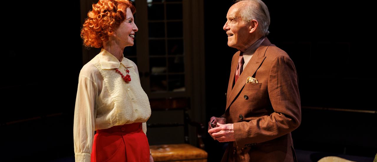 A woman in a cream-coloured blouse and red skirt stands on stage, smiling. She has curly red hair and a red jewelled necklace, and is looking at a man in a brown suite jacket with slick-backed grey hair. He is smiling warmly too. Behind them is a small brown stool and a door with glass windows.