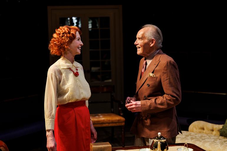 A woman in a cream-coloured blouse and red skirt stands on stage, smiling. She has curly red hair and a red jewelled necklace, and is looking at a man in a brown suite jacket with slick-backed grey hair. He is smiling warmly too. Behind them is a small brown stool and a door with glass windows.