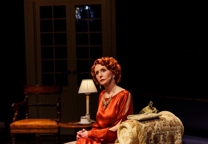 An older woman with red curly hair sits on a floral-patterned chaise longue. She wears a flowing, floor-length read dress and a large gold necklace and eaarings. Her head is cocked to one side, she looks distant and forlorn. Behidn her is a brown leather chair and a set of doors.