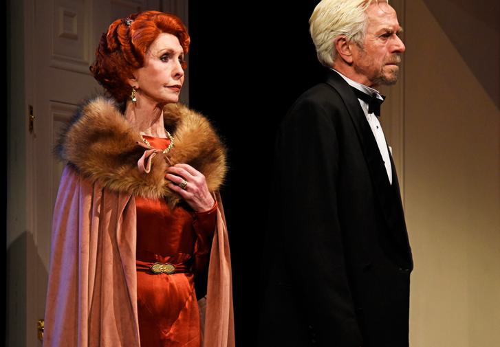 An elegant woman with curly red hair, wearing a rust-coloured cloak with a fur collar over a red satin dress, stands behind a man with white hair and beard, wearing a black dinner jacket. Both look to the right with serious expressions.