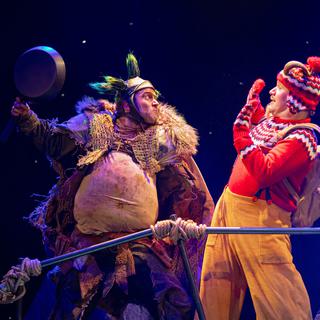 On the left, the Troll brandishes a frying pan towards Middle Goat, who holds his hands in front of his face in fright. The Troll has green tufts of hair and a big white stomach protruding from his tatty cothes. Middle Goat has a red and white Christmas jumper, with matching mittens and a woolly hat with a red pompom, through which stick his curly brown horns.