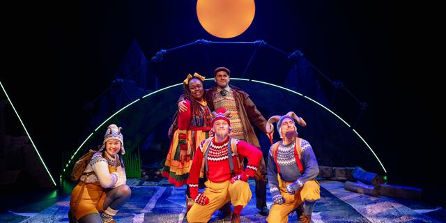 The three Goats, played by actors wearing bright Christmas jumpers and yellow trousers or dungarees, with horns on their woolly hats, kneel at the front of the stage looking forward and smiling. Behind them are standing Bo Frilly in a brightly striped dress and her Dad who's wearing a Fair Isle jumper and brown coat. An orange moon hovers above them.