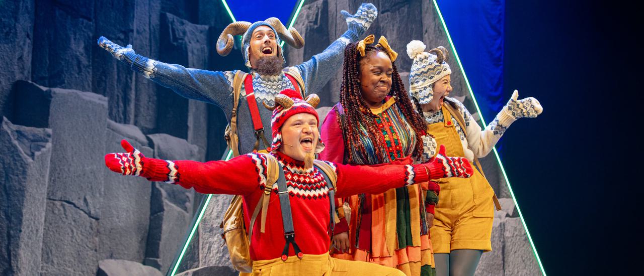 The three Goats, played by actors wearing bright Christmas jumpers and yellow trousers or dungarees, with horns on their woolly hats, are smiling broadly with their arms stretched out. They're surrounding Bo Frilly, who has long dark braids with yellow bows in her hair and wearing a brightly striped dress.