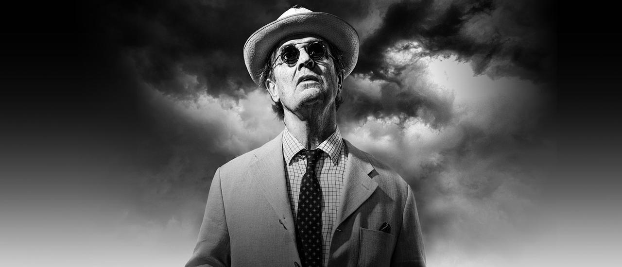 A black and white photograph of Rupert Everett in dark sunglasses, a trilby hat and a light coloured suit jacket with a fine-checked shirt and dark patterned tie. behind him is a backdrop of dark swirling clouds fading to a bright white at the bottom of the image.