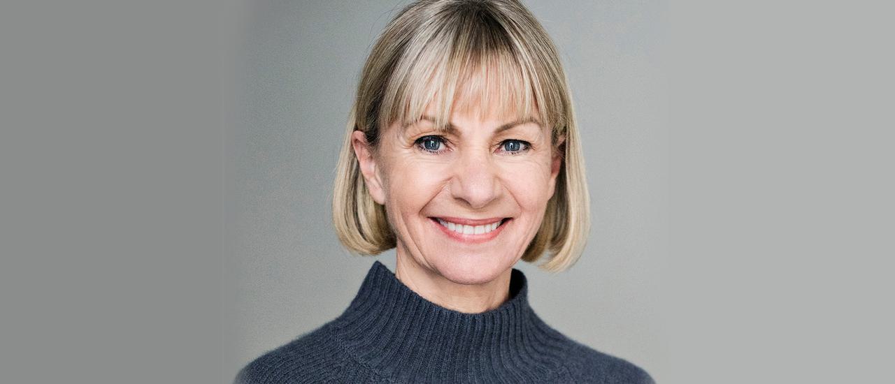 Kate Mosse smiles at the camera. She's wearing a grey high necked sweater. The background is a light grey.