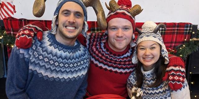 Three people in colourful clothing (woolly jumpers, braces, gloves and hat) with curly goat horns attached, cuddled together smiling at the camera.