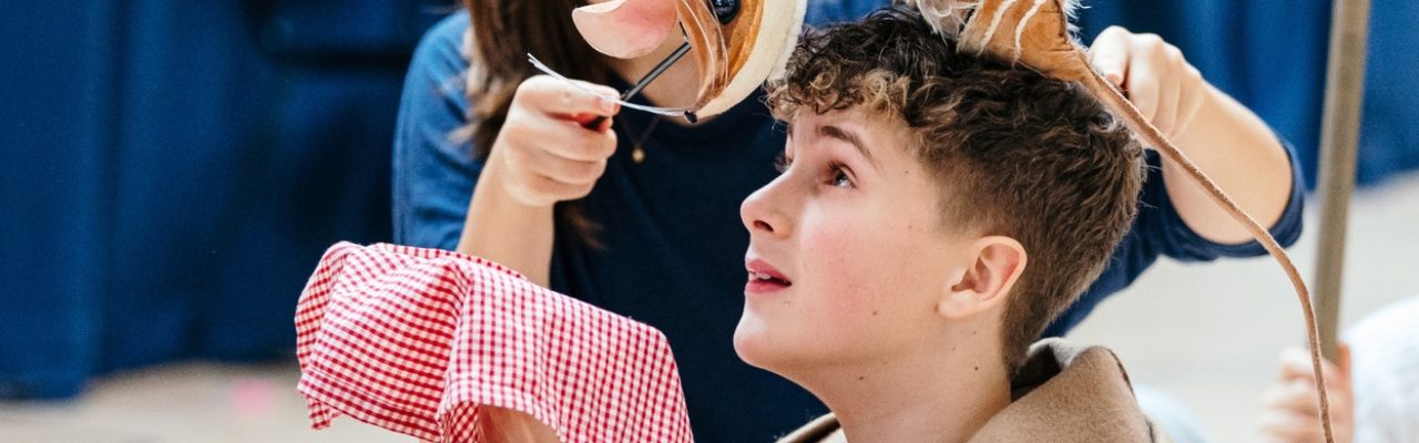 A young boy in a tan trench coat is sat on the floor with his legs crossed. He is holding a red gingham cloth out in front of his face. Next to him, a girl is operating a small mouse puppet, which is resting on his head. He is looking up at the mouse.