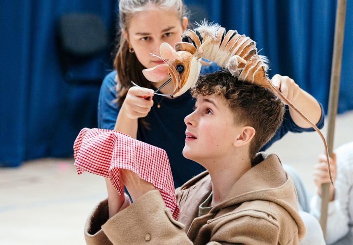 A young boy in a tan trench coat is sat on the floor with his legs crossed. He is holding a red gingham cloth out in front of his face. Next to him, a girl is operating a small mouse puppet, which is resting on his head. He is looking up at the mouse.