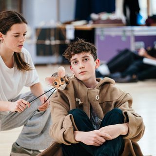 A young boy in a tan trench coat is sat on the floor with his knees pulled up to his chest. Next to him, a girl is operating a small mouse puppet, which is resting on his shoulder.