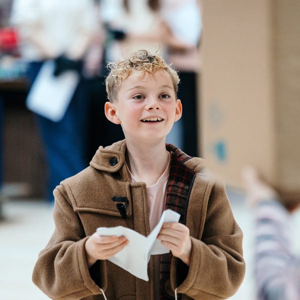 A young boy in a brown trench coat smiles gently. He is holding a folded piece of paper in his hands.