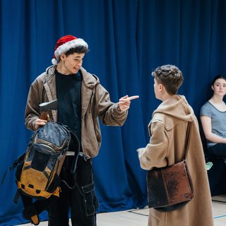 Two young performers are engaging in conversation. A taller boy in a brown jacket and a red Santa hat is holding a large backpack and an axe. He is pointing at a short boy in a beige trench coat with a worn leather satchel over his shoulder.
