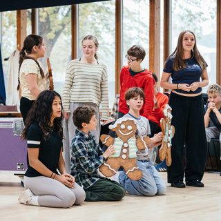 A group of young performers are in a rehearsal room. Four are standing and in front three are kneeling. Two of the performers are holding large decorated gingerbread men.