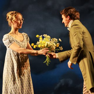 A young man in a brown Regency frock coat hands a bunch of flowers to a young woman wearing a paisley-patterned Regency dress. Her blonde hair is elegantly dressed and she is speaking to the man as she accepts the flowers.