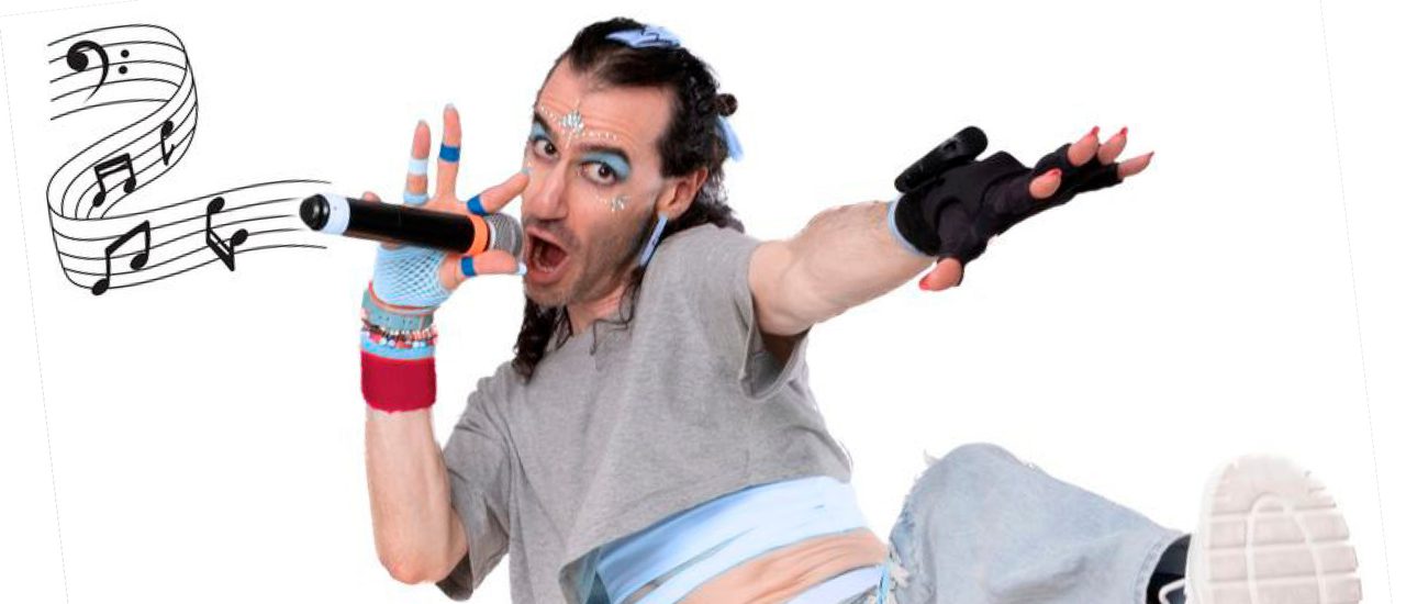 A performer wearing distressed light blue jeans, a cropped grey t-shirt, black fingerless gloves and chunky black boots stands on one leg, with the other leg kicking out in front of them. They look at the camera and speak into a microphone, with a graphic of musical notes coming out of it.
