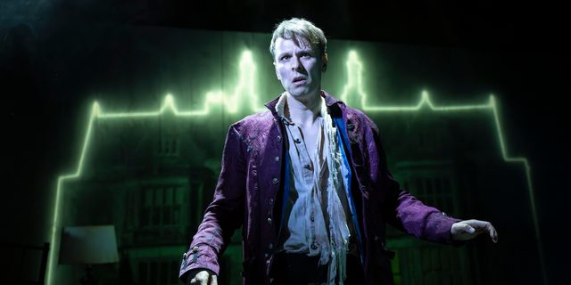 A young man stands in front of an old manor house, which is illuminated around the edge with a light green neon light. He wears a ragged white shirt with half of the buttons undone and a worn, deep purple tailcoat. He looks tense and concerned.