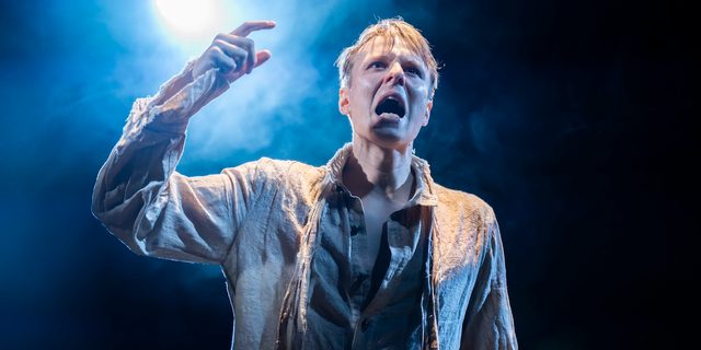 A young man is photographed dramatically from below with smoke surrounding his head. He is shouting and pointing with one hand. He wears a ragged off-white shirt with some of the buttons undone. A single blue spotlight illuminates the dark background and there is a light covering of smoke all around him.