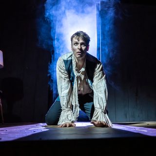 A young man kneels on all floors, looking up with a scared, tense expression. He wears a ragged off-white shirt with a trailing neck tie, blue waistcoat with the buttons undone and dark trousers. The background behind him is dark, but a doorway lets in an ominous wash of blue and purple lighting and smoke. There is a single lamp on a wooden stool to the left.
