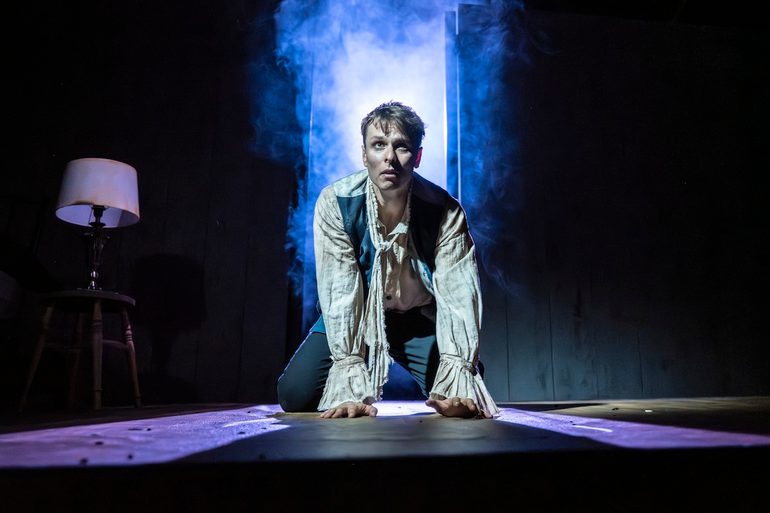 A young man kneels on all floors, looking up with a scared, tense expression. He wears a ragged off-white shirt with a trailing neck tie, blue waistcoat with the buttons undone and dark trousers. The background behind him is dark, but a doorway lets in an ominous wash of blue and purple lighting and smoke. There is a single lamp on a wooden stool to the left.