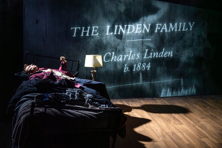 A young man lies on a single bed with a metal frame and dark bedding, with a lamp beside it. The man appears to be asleep. On the wooden backdrop, white text is projected, that reads: 'The Linden Family | Charles Linden b. 1884'. The bed and lamp are lit in white so that it casts a shadow on the wooden floor.